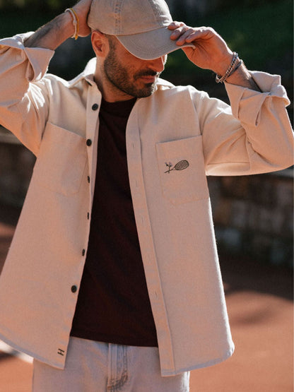 Man wearing a beige jacket with a logo, black shirt, and light-colored pants outdoors.
